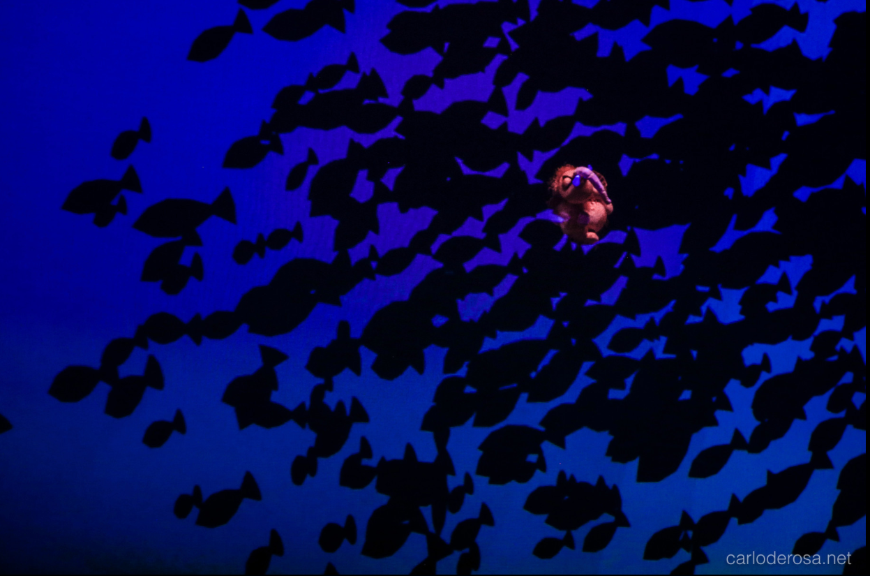 Moti traverse un décor sous-marin d’un bleu intense, au milieu des silhouettes de poissons.