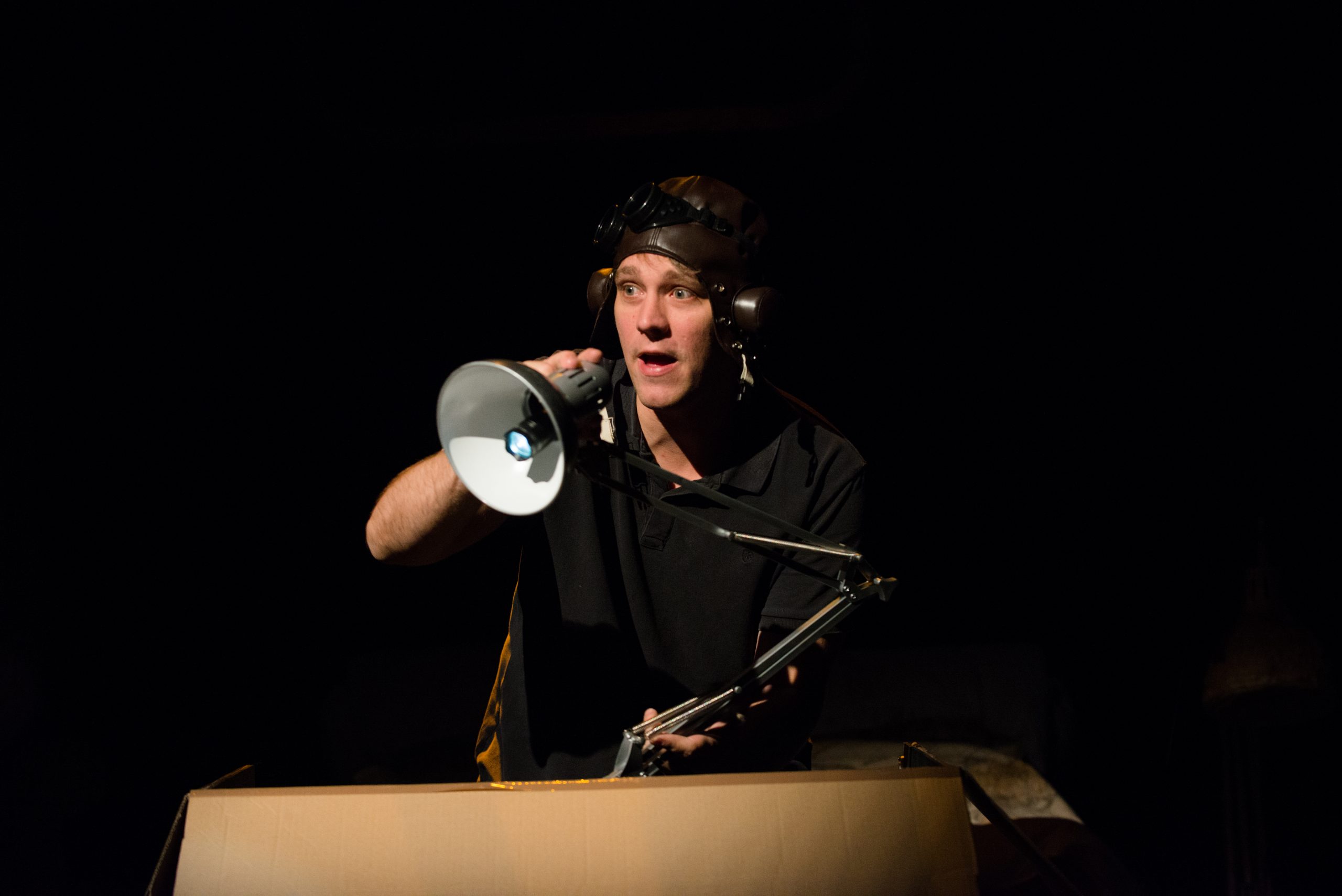 Le comédien coiffé d’un casque d’aviateur joue avec une lampe.